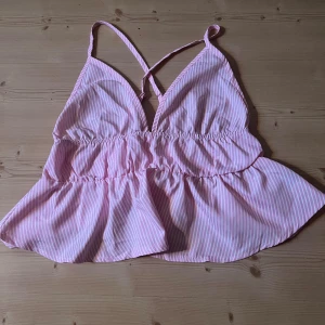 Rosa randig linnetopp med volang - Supersöt rosa och vit randig linnetopp med volangkant och smala axelband som korsas i ryggen. Toppen har justerbara band och en luftig, flowy passform.🥰