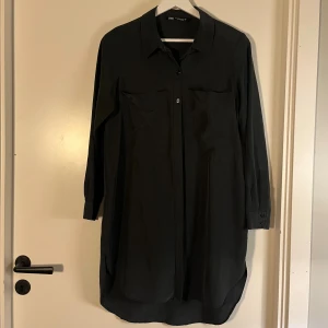 Svart skjortklänning från Zara XS - Svart skjortklänning från Zara i storlek XS i modal. Klänningen har klassisk krage, knappar framtill och två bröstfickor. Långärmad med knapp vid ärmslut. Perfekt för en clean och stilren look.