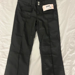 Svarta bootcut-byxor från Nelly - Svarta bootcut-byxor från Nelly med låg midja och snygga fickdetaljer framtill. Byxorna har två knappar i midjan och är tillverkade i ett slätt material som ger en cool look. Perfekt för dig som gillar trendiga silhuetter och detaljer.