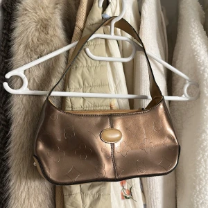 Bronzefärgad axelväska med mönster - Snygg bronzefärgad axelväska med präglade blommor och geometriska former. Väskan har ett beige knäppspänne framtill och dragkedja upptill. Insidan är ljusbeige med flera fack och dragkedja för smidig organisering. Perfekt storlek för det viktigaste.