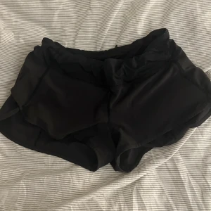 Svarta träningsshorts från Lululemon - Säljer ett par svarta träningsshorts från Lululemon med klassisk logga på sidan. Shortsen har loose passform, elastisk midja och är gjorda i ett lätt syntetmaterial som andas. Perfekta för gymmet eller löprundan, riktigt sköna och stilrena. Djur i hemmet,bra skick!!