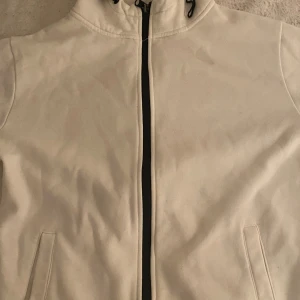 Vit vindjacka från Smog med huva - Vit vindjacka från Smog i slim fit-modell. Jackan har svart dragkedja framtill, justerbar huva med snörning och två fickor på sidorna. Perfekt för dig som gillar stilren och enkel design.