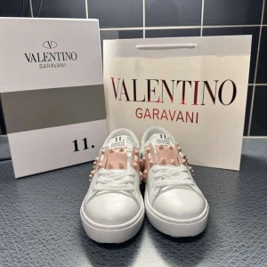 Valentino Garavani Rockstud sneakers - Säljer ett par vita Valentino Garavani Rockstud sneakers med ljusrosa panel och ikoniska nitar. Skorna har rund tå, platt sula och snörning. Tillverkade i skinn med coola detaljer som ger en lyxig vibe. Perfekt för dig som gillar statement pieces och designer.