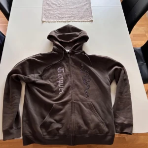 Brun hoodie med tryck och dragkedja - Säljer en brun hoodie från Trapper av en orm och logga på framsidan. Tröjan har huva, långa ärmar och praktisk dragkedja framtill. 