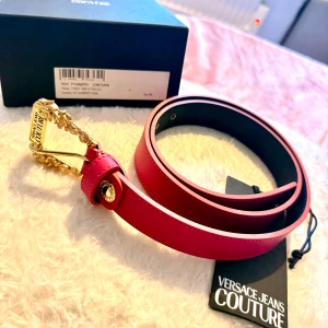 Rosa skärp från Versace Jeans Couture - Säljer ett snyggt rosa bälte från Versace Jeans Couture med exklusivt guldfärgat spänne och dekorativa detaljer. Bältet är tillverkat i skinn och har en stilren, slät design med svarta insidor. Perfekt accessoar för att lyfta din outfit.