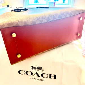 Brun och vinröd handväska från Coach - Snygg handväska från Coach i klassisk boxig form med bruna och vinröda paneler. Väskan har Coachs ikoniska mönster, guldfärgade detaljer och både handtag och axelrem. Materialet är skinn och canvas, perfekt för dig som gillar stilrena accessoarer med lyxig känsla.