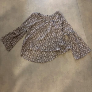 Mönstrad blus från Mon Row - Säljer en luftig blus från Mon Row i storlek L. Blusen har ett brunt och vitt geometriskt mönster, vida långa ärmar och en avslappnad passform. Perfekt för dig som gillar boho-vibbar och unika prints.