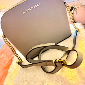 Grå axelväska från Michael Kors - Snygg grå axelväska från Michael Kors med guldfärgade kedjedetaljer och justerbar axelrem. Väskan har en stilren, rundad form och är tillverkad i skinn med logotyp framtill. Invändigt finns ett fack med dragkedja och plats för småsaker.