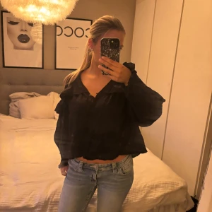 Svart blus med volang och knappar - Säljer en svart blus med volangdetaljer och knappar framtill. Blusen har långärmade, lite puffiga ärmar och en croppad passform som ger en cool siluett. Materialet ser ut att vara lätt och luftigt, perfekt för att styla med jeans eller kjol.