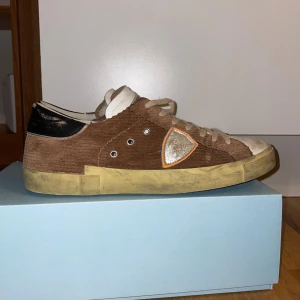 Philippe Model bruna sneakers i manchester - Säljer ett par snygga Philippe Model sneakers i brun manchester med vita detaljer och beige snörning. Skorna har en rund tå, platt sula och en silverfärgad sköld med orange kant på sidan. Svart hälparti och slitna detaljer för en cool look.