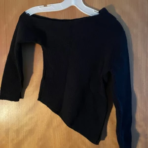 Svart stickad offshoulder topp - Svart stickad topp med offshoulder-design och lång ärm. Asymmetrisk nederkant ger en cool och unik look. Perfekt för dig som gillar att sticka ut med stilrena detaljer och sköna material.