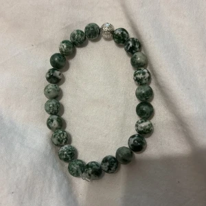 Grönt pärlarmband med silverdetalj - Stilrent armband med runda gröna pärlor i marmorerat mönster och en glittrig silverfärgad pärla som detalj. Elastiskt band gör det enkelt att ta på och av. Perfekt accessoar för att lyfta din outfit med en cool touch.