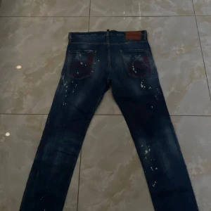 Desquared  jeans med slitningar och färgstänk - Mörkblå jeans från desquared med coola slitningar och vita färgstänk på både fram- och baksida. Klassisk femficksmodell med raka ben och snygga detaljer på bakfickorna. Tillverkade i 100% denim med italiensk design, perfekt för dig som gillar streetstyle.