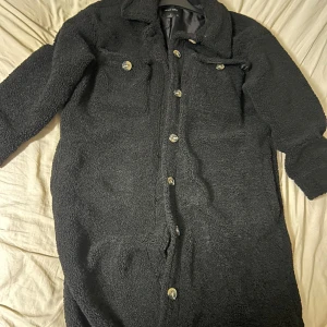 Svart teddyjacka från Brave Soul - Mysig svart teddyjacka från Brave Soul i storlek M. Jackan har en längre modell, krage, två bröstfickor med knappar och stängs med silvriga knappar framtill. Materialet är fluffigt och ger en cool oversized känsla. Perfekt för kalla dagar när du vill vara både varm och snygg.