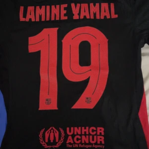 FC Barcelona Lamine Yamal tröja Nike - Säljer en svart FC Barcelona fotbollströja med Lamine Yamal och nummer 19 på ryggen. Tröjan har röda detaljer, UNHCR och Spotify-logga, samt klubbmärke på bröstet. Kortärmad modell i lätt och ventilerande material, perfekt för match eller träning.