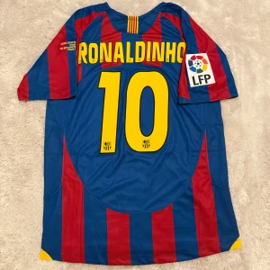 FC Barcelona Ronaldinho #10 tröja Nike - Klassisk FC Barcelona fotbollströja med Ronaldinho och nummer 10 på ryggen. Tröjan är blå och röd med gula detaljer, randigt mönster och officiella klubbmärken samt LFP-patch på ärmen. Tillverkad i lätt och ventilerande polyester.