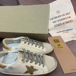 Golden Goose sneakers med glitterstjärna - Snygga vita sneakers från Golden Goose med grå snörning, guldiga detaljer på hälen och tungan samt en glittrig stjärna på sidan. Skorna har en platt sula med slitna effekter och är tillverkade i skinn och mocka. Perfekt för dig som gillar unik design och coola detaljer.