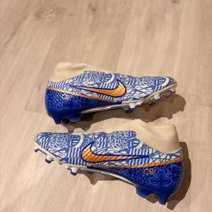 Nike Mercurial CR7 blå fotbollsskor - Säljer ett par Nike Mercurial CR7 fotbollsskor med blått och vitt grafiskt mönster, guldiga detaljer och logga. Skorna har platt sula med dobbar för gräsplan och konstgräs. en strumpliknande överdel i syntetmaterial för tight passform. Snygg design med CR7-tryck på sidan och Mercurial-text bak.