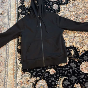Svart hoodie från Burberry med dragkedja - Svart hoodie från Burberry med hel dragkedja och lhuva med snörning. Insidan av lhuvan har klassiskt rutigt Burberry-mönster i svart, vitt och grått. Tröjan har två fickor framtill och är tillverkad i bomull och polyester. Perfekt för en stilren och avslappnad look. Vill sälja den för att den passar inte mig. Funkar för båda dem som har storlek M och L