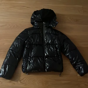 Svart pufferjacka med huva från Nelly - Säljer en svart, glansig pufferjacka från NA-KD med huva och dragkedja framtill. Jackan har quiltad design, långa ärmar och justerbar nederkant med snörning. Perfekt för kalla dagar och riktigt snygg till streetwear-stilen.