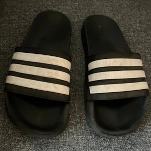 Svarta Adidas adilette sandaler med vita ränder - Säljer ett par klassiska svarta Adidas sandaler med tre vita ränder över foten. Perfekt för chill dagar eller att glida runt hemma. De har en platt sula och är gjorda i syntetmaterial för enkel rengöring.