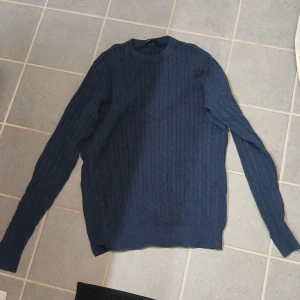 Mörkblå stickad tröja med kabelmönster - Snygg mörkblå stickad tröja med klassiskt kabelmönster. Tröjan har rund hals och långa ärmar, perfekt för att hålla värmen under kyliga dagar. Passar dig som gillar stilrena och tidlösa plagg med lite extra struktur.