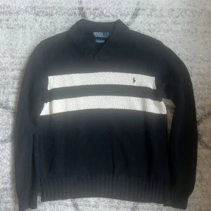 Svart tröja med vita ränder Polo Ralph Lauren - Klassisk svart tröja från Polo Ralph Lauren med två breda vita ränder över bröstet och broderad logga. Tröjan har v-ringad krage och är ribbstickad vid ärmslut och nederkant. Tillverkad i 100% bomull och har lång ärm.