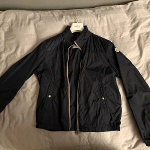 Mörkblå vindjacka från Moncler - Säljer en mörkblå vindjacka från Moncler med klassisk logotyp på ärmen. Jackan har ståkrage, dragkedja med rödvit kant, två fickor med tryckknappar och elastisk nederkant. Materialet är lätt och syntetiskt, perfekt för blåsiga dagar. Långärmad och stilren design.