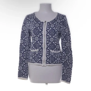 Blåvit stickad kofta från RoseBud - Blåvit stickad kofta från RoseBud med klassiskt nordiskt mönster. Koftan har rund hals, två fickor framtill och stängs med dragkedja. Perfekt för kalla dagar och ger en mysig vintervibe.