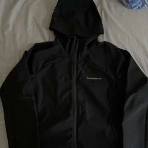 Svart vindjacka Peak Performance - Svart vindjacka från Peak Performance med huva och hel dragkedja framtill. Jackan har en clean look med diskret logga på bröstet och praktiska fickor med dragkedja. Perfekt för dig som gillar sportig och stilren design.