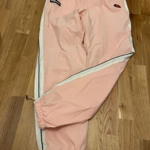 Rosa byxor (tracksuit) från Ellesse, strl 14/L - Snygga ljusrosa byxor tracksuit style från Ellesse med vita sidopaneler och svarta detaljer. Byxorna har elastisk midja, meshfoder och snörning vid benslut för extra komfort. Perfekt för dig som gillar sportig stil och vill sticka ut med färg.