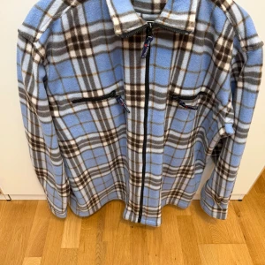 Blårutig fleecejacka Canada Highway XXL - Blårutig fleecejacka från Canada Highway i storlek XXL. Jackan har dragkedja framtill, två bröstfickor med dragkedja och klassisk krage. Mönstret är rutigt i blått, brunt och vitt, och materialet är mjuk fleece som är perfekt för kyliga dagar.