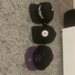 3 kepsar - Säljer tre snygga kepsar! En svart Norrlands keps med vit text, en svart New York Yankees keps med rund vit och röd logga, samt en svart keps med lila skärm och lila NY-brodyr. På New York Yankees kepsen finns de en liten orange fläk ovanför märket som förmodligen går bort om man tvättar den vilket jag inte provat. 