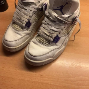 Nike Air Jordan 4 Retro vit/lila - Nike Air Jordan 4 Retro sneakers i vit skinn med lila detaljer på snörningen och Jumpman-logga på plösen. Klassisk meshpanel på sidorna och platt sula med synlig Air-enhet. Rund tå och snörning för en ikonisk look.