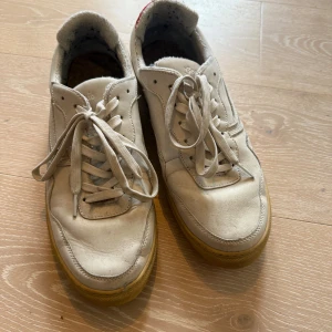 Genesis sneakers med röd detalj - Säljer ett par vita sneakers från Genesis med coola röda detaljer i fiskfjällsmönster på hälen. Skorna har snörning, platt sula och är tillverkade i skinn med inslag av textil. Snygg kontrast mellan den ljusa ovandelen och den röda baksidan. Perfekt för dig som gillar unika sneakers.