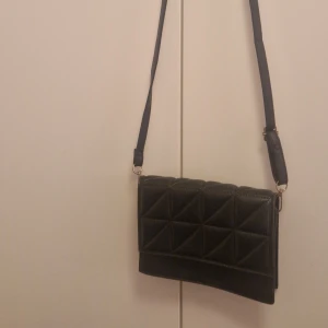 Svart quiltad axelväska i skinnimitation - Stilren svart axelväska med quiltad framsida i geometriskt mönster. Väskan har justerbar axelrem och stängs med magnetlås. Guldiga metalldetaljer och rektangulär form. Perfekt för att bära det viktigaste och passar till många outfits.