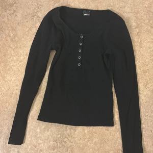 Svart ribbad topp från Gina Tricot - Svart långärmad topp från Gina Tricot i ribbad bomull med rundad hals och fem dekorativa knappar framtill. Snygg och enkel design som funkar till många olika stilar. Perfekt för dig som gillar stilrena och bekväma plagg.