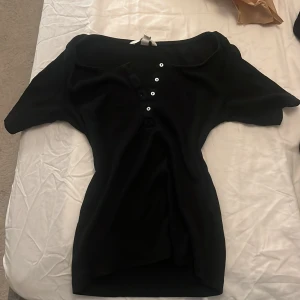 Svart ribbad topp från H&M med knappar 2000s elena gilbert - Svart ribbad topp från H&M i storlek S. Toppen har korta ärmar, rundad hals och dekorativa vita knappar framtill. Materialet är mjukt och stretchigt, perfekt för en chill och stilren look.