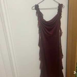 Vinröd glittrig långklänning - Vinröd långklänning med glittriga detaljer och volanger längs axelbanden och sidorna. Klänningen har en draperad urringning och är ärmlös, vilket ger en elegant och festlig vibe. Materialet är lätt och skimrande, perfekt för att sticka ut på bal eller fest.