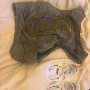 Grå Osprey Duro 1.5 löparväst - Säljer en grå Osprey Duro 1.5 löparväst med orange detaljer och flera smarta fack. Västen har dragkedja framtill och elastiska snören för extra förvaring. Två mjuka 500ml flaskor medföljer, perfekt för längre löpturer eller äventyr.