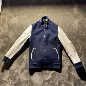 Blå och vit bomberjacka Zadig & Voltaire - Snygg blå bomberjacka från Zadig & Voltaire med vita ärmar och vita detaljer vid fickorna. Jackan har ribbade muddar, knappar framtill och en cool dragkedjedetalj vid kragen. Materialmix med ull och skinnkänsla på ärmarna. Perfekt för dig som vill sticka ut.