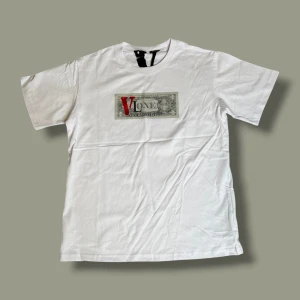 Vit VLONE t-shirt med dollartryck - Säljer en vit t-shirt från VLONE i 100% bomull, med stort dollartryck och röd V-logga på bröstet. På ryggen finns en stor V med dollar-motiv. Klassisk rund hals och korta ärmar. Perfekt för dig som gillar streetwear och unika prints.
