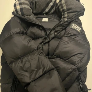 Burberry Jacka - Svart pufferjacka från Burberry med stickad luva och logotyp på axeln. Jackan har dragkedja och knappar framtill, två sidofickor med dragkedja och quiltad design. Materialet är polyester och dun, perfekt för kalla dagar. Snygg och stilren look med klassisk Burberry-vibe. I storlek M och kan även passa S