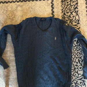 Blå kabelstickad tröja Polo Ralph Lauren - Kabelstickad blå tröja från Polo Ralph Lauren med v-ringning och broderad logga på bröstet. Tröjan har lång ärm och klassisk passform, perfekt för dig som gillar en stilren och tidlös look. Gammal tröja så den är ner tvättad till en S