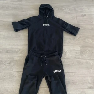 Svart hoodie från Hoodrich med tryck - Svart hoodie från Hoodrich med vit logga på bröstet, huva med snörning och stor magficka. Tröjan är i mjuk bomullsmix och har klassisk passform med långa ärmar. Perfekt för en chill streetwear-look.