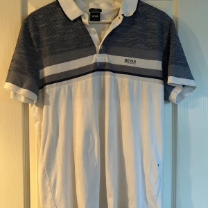 Vit och blå pikétröja från Hugo Boss - Snygg pikétröja från Hugo Boss i slim fit med klassisk krage. Tröjan är vit med blå partier upptill och har subtila ränder samt logga på bröstet. Tillverkad i lyxig bomull och har korta ärmar. Perfekt för en stilren och fräsch look.