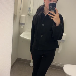 Svart kort kappa - Säljer en skit snygg svart, kort kappa stora framfickor och knappar framtill. Kappan har en klassisk krage och långärmade ärmar. Passar dig som vill ha en clean look, kappan är inprincip helt ny bara använd ett fåtal gånger men faller inte riktigt mig i smaken så hoppas någon annan som gillar denna stil mer kan ta rätt på den så slipper den hänga i garderoben 