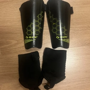 Svarta benskydd från SOC - Säljer ett par svarta benskydd från SOC med gulgrönt grafiskt mönster. Skydden har en böjd form för att passa smalbenet och kommer med svarta elastiska strumpor för extra komfort. Perfekt för fotbollsträning eller match.