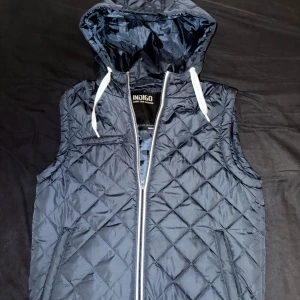 Mörkblå quiltad dunväst med huva - Quiltad mörkblå dunväst från Indigo med huva och vita dragsnören. Västen har dragkedja framtill, två fickor med dragkedja och en bröstficka. Perfekt för lager-på-lager och snygg till streetwear.