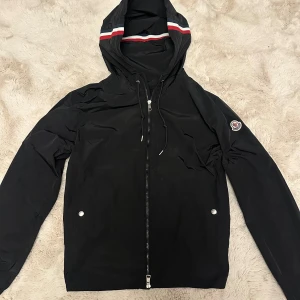 Svart Moncler vindjacka L - Svart vindjacka från Moncler med huva och coola röd, vit och blå ränder på kanten. Jackan har dragkedja framtill, två fickor med tryckknappar och Moncler-logga på ärmen. Materialet är lätt och syntetiskt, perfekt för blåsiga dagar.